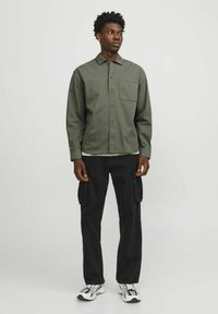 Oliven grøn skjorte med knapper og brystlomme, sorte cargo-bukser med sidelommer, og hvide sneakers med grå detaljer.