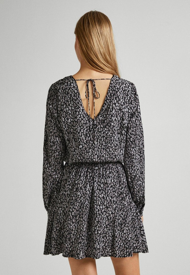 Femme aux longs cheveux blonds portant une robe imprimée léopard noire et grise avec des manches longues et un dos ouvert noué avec un nœud.