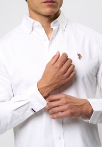 Biała koszula zapinana na guziki z małym haftowanym wzorem na piersi, długimi rękawami z mankietami na guziki oraz kołnierzykiem.