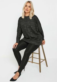 Czarny, fakturowany oversize sweter z okrągłym dekoltem, w połączeniu z błyszczącymi czarnymi legginsami. Modelka siedzi na drewnianym stołku, nosząc czarne szpilki.