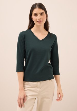 Camisa verde oscuro de manga larga con escote en V, fabricada en una tela suave. Presenta un diseño ajustado y una textura lisa, combinada con pantalones beige.