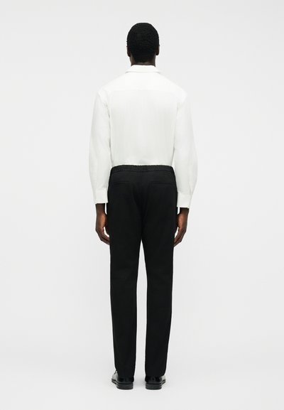 Άνδρας με την πλάτη, φοράει λευκό πουκάμισο με μακριά μανίκια, μαύρο παντελόνι με πίσω τσέπες και μαύρα παπούτσια, πάνω σε απλό λευκό φόντο.