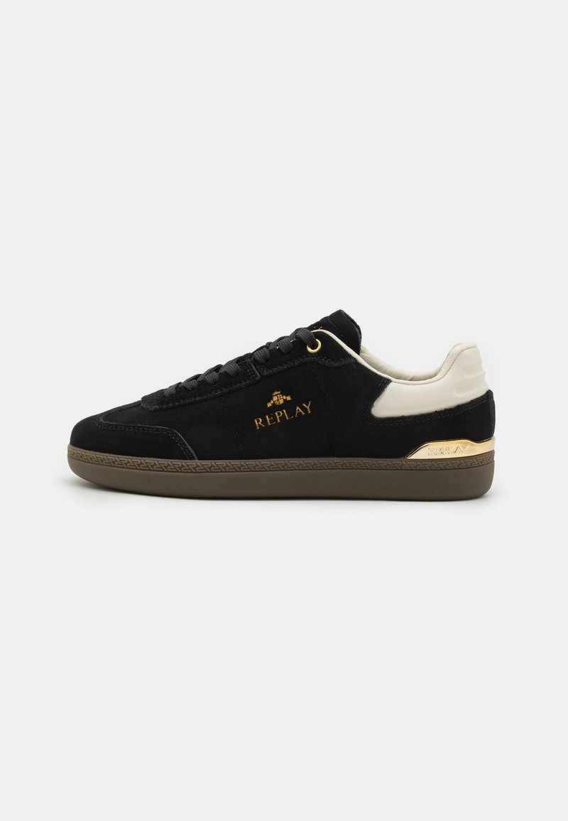 Black suede sneaker with a cream leather accent and gold hardware. Features a textured brown rubber sole and embossed logo detailing.