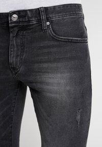 Svarta denimjeans med en urtvättad yta, som har en knappstängning i midjan och en liten sliten patch på låret.