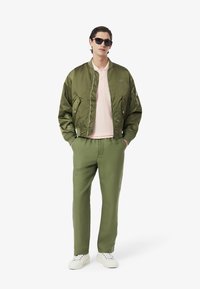 Grön bomberjacka, rosa pikétröja, gröna byxor och vita sneakers. Funktioner inkluderar dragkedja fram, sidofickor och logotypdetalj på jackan.