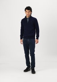 Mornaricky pulover s polu-zip ovratom, pletena tekstura, uparen s tamnoplavimfarmerkama i crnim tenisicama, stoji nasuprot bijeloj pozadini.