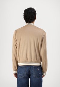 Tan bomberjakke med glatt tekstur, ribbede mansjetter og stripet liv. Båret med blå slitte jeans, sett fra ryggen.