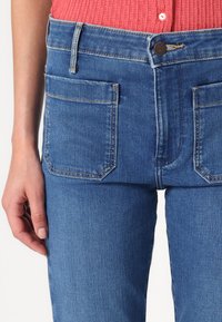 Jean en denim bleu avec une coupe droite, comportant des poches avant et arrière, ainsi qu'une fermeture à bouton. Des détails de couture discrets sont visibles.