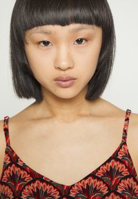 Modelo a usar uma peça de vestuário com padrão floral vermelho e alças finas. Apresenta cabelo preto liso e maquilhagem mínima, com um fundo neutro.