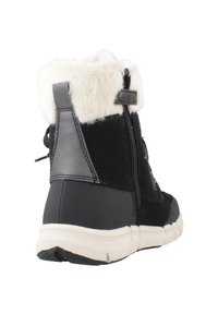 Botín negro de invierno con puño de piel blanca, cremallera lateral, tacón texturizado y suela de goma blanca, visto ligeramente desde atrás.