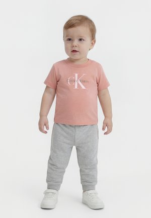 Smårolling iført en rosa Calvin Klein t-skjorte, grå joggebukser og hvite joggesko, stående på hvit bakgrunn.