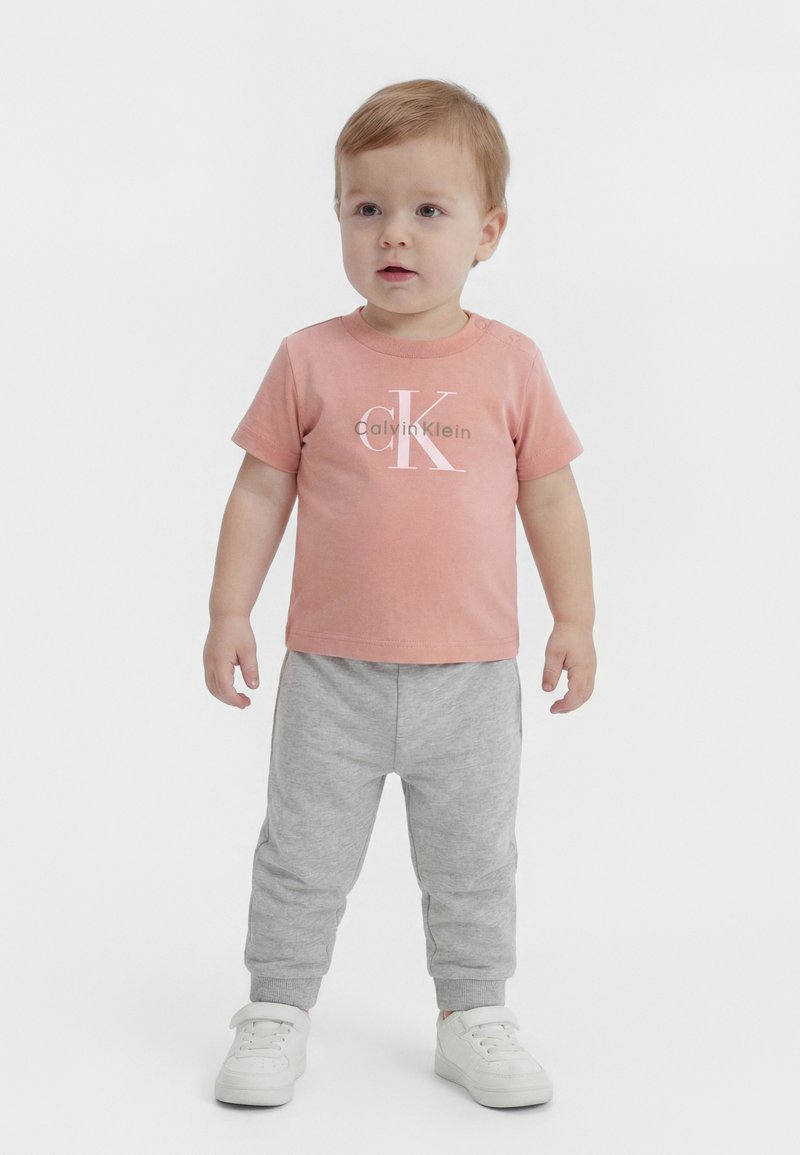 Tout-petit portant un t-shirt rose Calvin Klein, un pantalon de survêtement gris et des baskets blanches, debout sur un fond blanc.