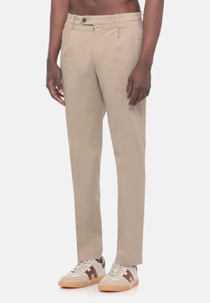 Uomo con pantaloni beige a gamba dritta e sneakers bianche con logo marrone "H", in piedi su uno sfondo bianco.