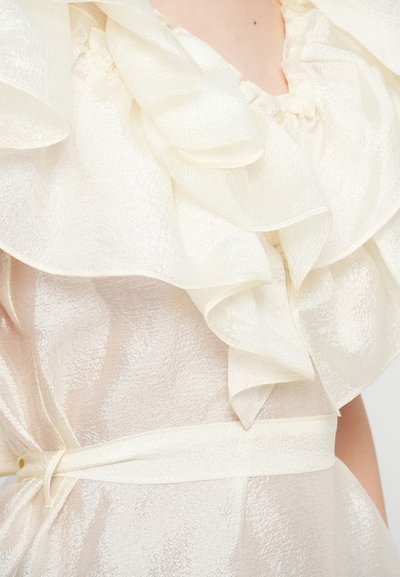 Mirdzoša krēmkrāsas auduma kleita ar kārtām un jostu ap vidukli, kas atklāj augšējo ķermeni un roku.