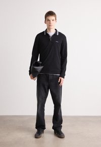 Polo preto de mangas longas com detalhes roxos, combinado com jeans escuros. O modelo segura uma clutch preta texturizada. Fundo neutro.