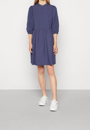 Femme portant une robe violette jusqu'aux genoux avec des manches bouffantes trois-quarts et des baskets blanches, se tenant devant un fond clair uni.