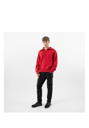 Mladý muž stojí, má na sobě červený svetr s krátkým zipem, černé kalhoty a černé tenisky na jednobarevném bílém pozadí.