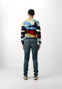 Suéter de punto multicolor con patrones abstractos; cuenta con mangas largas y cuello redondo. Combinado con jeans azules ajustados y zapatos casuales.