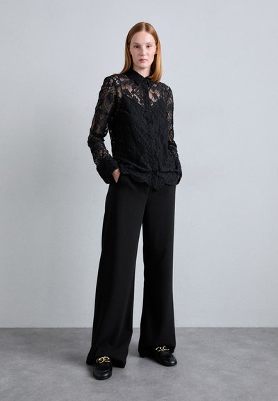 Μαύρη δαντελένια μπλούζα με μακριά μανίκια και γιακά, συνδυασμένη με μαύρο παντελόνι σε φαρδιά γραμμή. Λείος υφασμάτινος χαρακτήρας και μαύρα παπούτσια με χρυσές λεπτομέρειες.