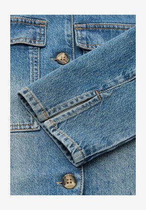 Veste en jean bleu clair avec fermeture à boutons, coutures visibles et deux poches poitrine. Présente un revers de manche plié avec une texture lisse.