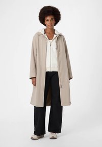 Cappotto impermeabile beige con cappuccio, bottoni a pressione e vestibilità comoda. Indossato sopra una giacca bianca con zip e pantaloni neri a gamba larga.