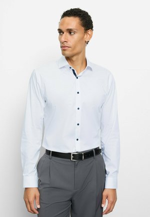 Langärmliges weißes Hemd mit marineblauen Knöpfen und Kragenakzenten, kombiniert mit grauer Hose und schwarzem Gürtel. Glatter Stoff und maßgeschneiderte Passform.