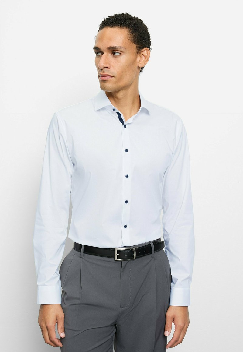 Langärmliges weißes Hemd mit marineblauen Knöpfen und Kragenakzenten, kombiniert mit grauer Hose und schwarzem Gürtel. Glatter Stoff und maßgeschneiderte Passform.