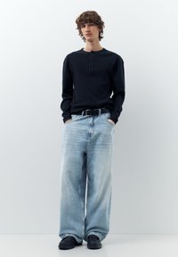 Ung person, der står op ad en ensfarvet væg iført en sort langærmet henley, lyse vide jeans, sort bælte og sorte sko.