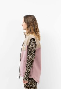Fleece-Weste in Beige und Pink mit Reißverschluss vorne, seitlichen Taschen und einem kleinen Logo-Patch. Getragen über einem langärmeligen Shirt mit Leopardenmuster.