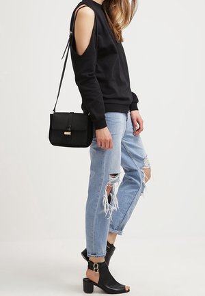 Svart sweatshirt med cold-shoulder-design, ljusblå slitna jeans och svarta ankelskots med spännde detaljer. Svart axelremsväska ingår.