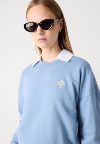Sweatshirt bleu clair avec un col côtelé, arborant un petit logo géométrique doré sur la poitrine. Lunettes de soleil noires style œil de chat, texture lisse.