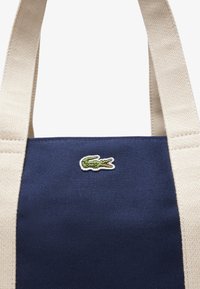 Sac fourre-tout en toile avec un corps bleu marine, des poignées crème et un logo de crocodile brodé vert. Design durable et rectangulaire avec un tissu texturé.
