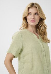 Blusa di lino verde chiaro con scollatura a V, maniche corte e chiusura frontale con bottoni. La texture è morbida e leggermente sgualcita.
