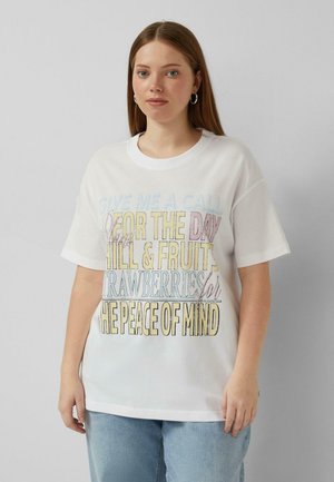 Junge Frau mit langen braunen Haaren, die Creolen trägt, ein weißes Grafik-T-Shirt mit pastellfarbenem Text und hellblaue Jeans vor einfachem Hintergrund.