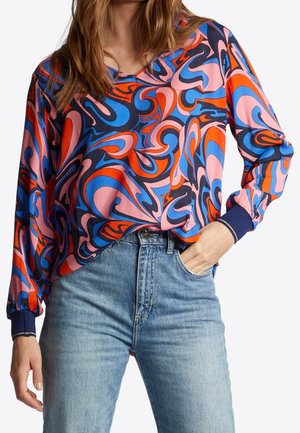 Mujer con una blusa de manga larga con un patrón abstracto de remolinos en rosa, azul, naranja y negro, combinada con vaqueros azul claro.
