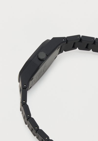 Πλάγια όψη μαύρου μεταλλικού ρολογιού με συνδεδεμένο μπρασελέ, εξαγωνική κορώνα και ορθογώνια θήκη σε λευκό φόντο.