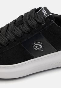 Svart mocka sneakers med tjocka svarta snören, en glansig svart logodetalj och en vit gummisula. Mjuk textur och låg skon design.