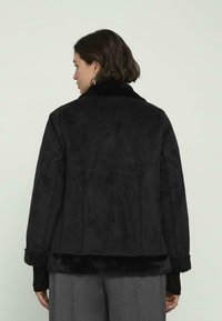 Donna con capelli corti e scuri che indossa una giacca nera foderata in pelliccia e pantaloni grigi, vista di spalle su uno sfondo chiaro e uniforme.