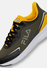 Vihreä oliivinvärinen urheilujalkine, jossa on mustia ja keltaisia yksityiskohtia, verkkomateriaali päällinen, paksu valkoinen pohja, varustettuna nauhoilla ja brändilogolla.