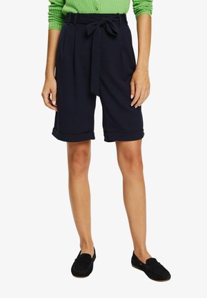 Femme portant un short noir taille haute longueur genou avec une ceinture nouée, ourlets retroussés, assorti à des mocassins noirs et un pull vert.