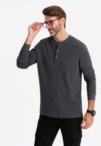 Mann, der schwarze runde Brillen anpasst, trägt ein dunkelgraues Henley-Shirt mit drei Knöpfen und schwarze Cargo-Hosen und steht vor einem weißen Hintergrund.