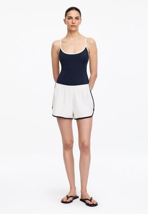 Vrouw staande met handen achter de rug, gekleed in een zwart tanktop, witte korte broeken met zwarte bies en zwarte sandalen op een witte achtergrond.