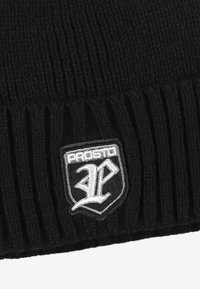 Czarna dzianinowa czapka z prążkowaną strukturą. Z przodu znajduje się biała naszywka z haftowanym logo na kontrastującym czarnym tle.