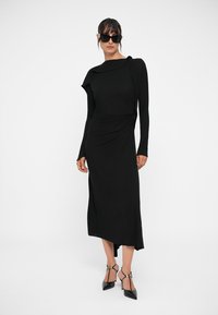 Juodas ilgas suknelė su drapiruotu dizainu, asimetrišku sijonu ir minkšta tekstūra. Suderinta su elegantiškomis juodomis stiletto kulniukais.