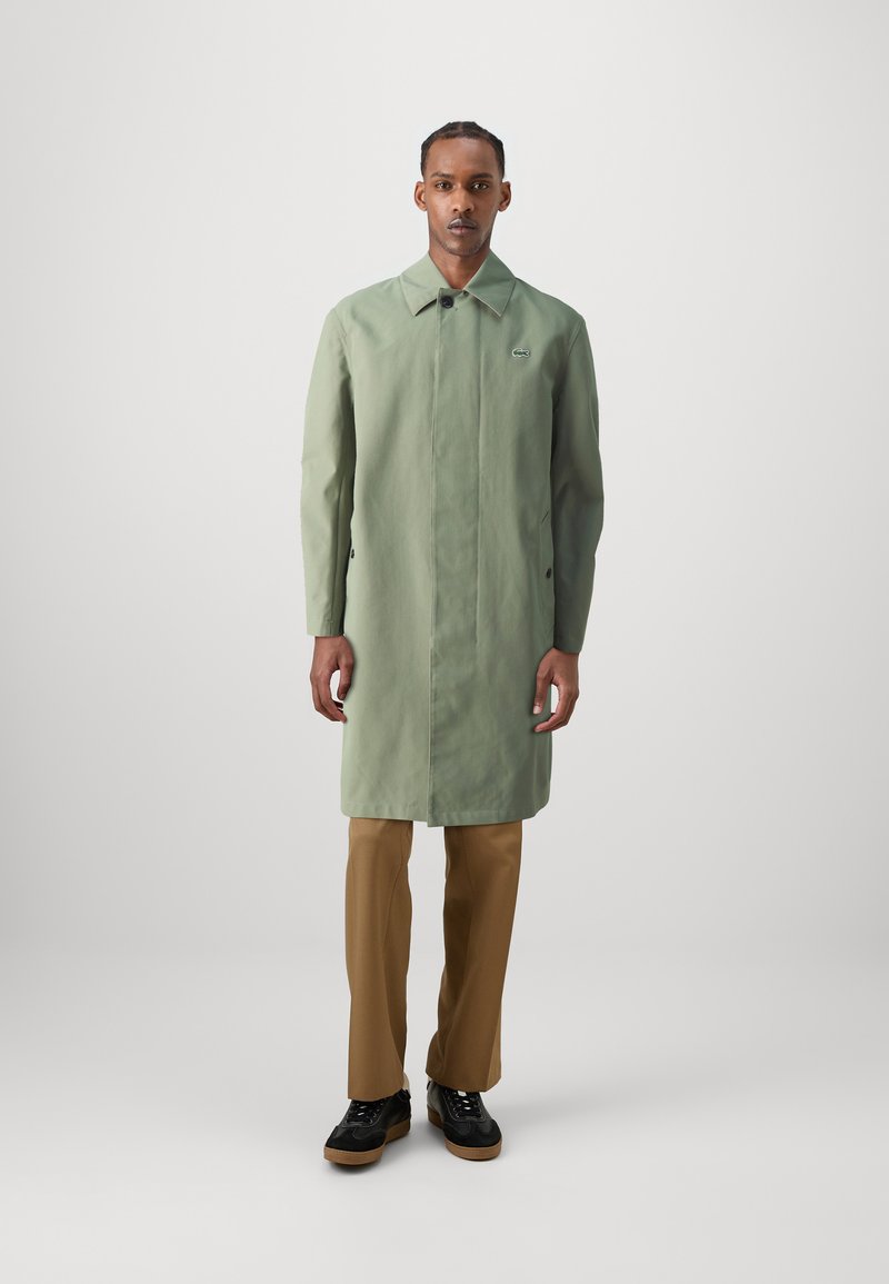 Cappotto verde chiaro con un design strutturato, caratterizzato da un colletto appuntito, maniche lunghe e tasche laterali. Indossato su pantaloni beige e scarpe nere.