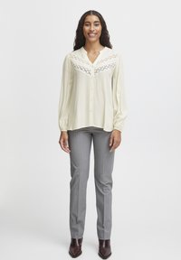 Cremefarvet bluse med blonde yoke-detalje, lange ærmer og knaplukning, parret med grå stribede bukser og brune ankelstøvler.