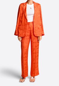 Helles oranges Zweiteiler-Anzug aus strukturiertem Material, bestehend aus einem gemusterten Blazer und geraden Hosen, kombiniert mit silbernen Sandalen.