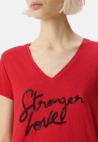 Camiseta de algodón roja con escote en V, que presenta un gráfico negro "Stronger Love" decorado con acentos de cuentas. Textura suave y mangas cortas.