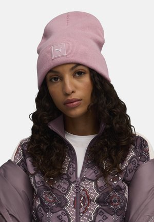 Beanie rosa in maglia con texture a coste e un'etichetta quadrata con il logo Puma. Abbinato a una giacca bomber viola con un motivo geometrico.