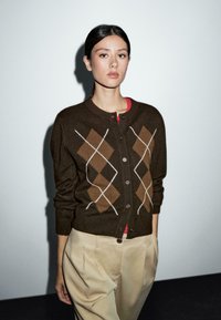 Junge Frau steht drinnen und trägt einen braunen Rautenmuster-Pullover, beige Hosen und ein rotes Hemd darunter, vor einer schlichten hellen Wand.